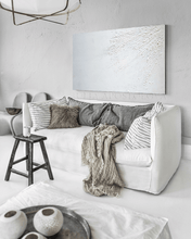 White couch with gray pillows and a blanket in a white room with a large white painting on the wall and a stool in front of the couch . The painting is minimalist  with rich white 3d relief textures and the whole room has wabi sabi decor.