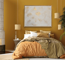 Cozy bedroom with mustard yellow walls, white gold minimalist painting, a bed with patterned bedding, and decorative elements, like lights and plans