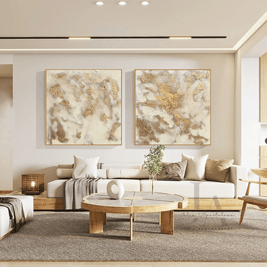Two abstract paintings with gold, and white colors in a living room with a white sofa , surrounded with pillows and a round wooden coffee table. The paintings are framed with gold floating frames, and featured metallic gold ,  textured abstract design, ivory and white colors and gloss resin finish provide a unique and eye-catching element for neutral decors.