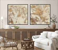 Two abstract paintings with gold, and white colors hanging over wooden cadenza in a living room with a white sofa , and a square wooden coffee table. The paintings are framed with black floating frames, and featured metallic gold , textured abstract design, ivory and white colors and gloss resin finish provide a unique and eye-catching element for neutral decors.