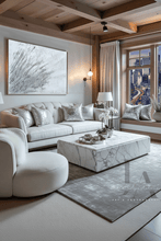 A luxury modern living room featuring a cream tufted sofa with silver glitter pillows, a matching round white armchair, a white marble coffee table, and a gray patterned rug. A large abstract white and silver wall art piece hangs above the sofa, set against a background of wood ceiling beams and a window overlooking a city skyline.