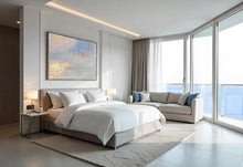 Modern bedroom with a bed, abstract seascape painting, sofa, and large windows with a view of water.