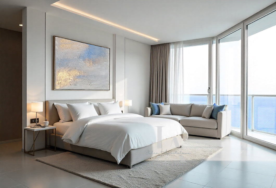 Modern bedroom with a bed, abstract seascape painting, sofa, and large windows with a view of water.