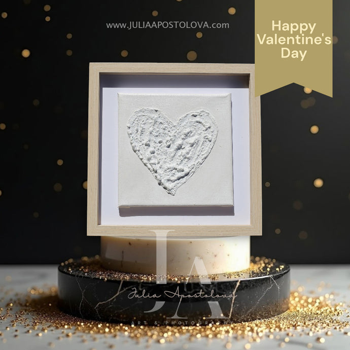 A minimalist white heart-shaped sculptured wall art painting on stretched canvas, framed with wooden shadow box frame and displayed on a marble stand with gold glitter around it. The background is dark , creating a dramatic contrast. Perfect art gift for Christmas, Valentine's Day, Birthday, Anniversary, and any occasion.