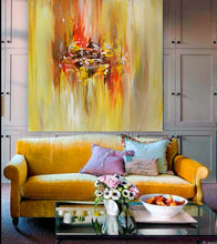 A large abstract canvas painting with vibrant yellow, orange, red and brown brushstrokes hangs on a gray paneled wall above a mustard yellow velvet sofa with decorative pillows and a glass coffee table in the foreground.