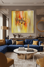 A large framed abstract painting with vibrant yellow, orange, and brown brushstrokes hangs on a beige wall above a blue sectional sofa in a modern living room setting.