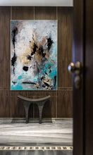 Abstract painting with black, white, gold, and teal paint on a canvas. The painting is hanging on a wall in a room with a wooden floor and a wooden bench.