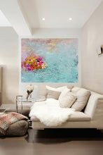 A large abstract canvas painting with turquoise, pink, and gold tones hangs on a wall above a modern beige sofa with white pillows and a white faux-fur throw in a bright, contemporary living room.