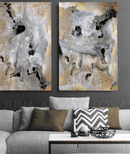A set of two contemporary abstract art paintings with gold, black, and gray colors, displayed above a sofa in a cozy living room setting.