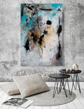 Abstract painting in shades of teal, gold, and black on a white canvas. The painting is hanging on a gray concrete wall above two black metal side tables with a gray rug and two pillows on the floor.